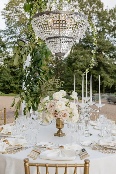 Wedding white table with luxurious classic-style serving with bouquet of roses in vintage vase under antique crystal chandelier on summer terrace of restaurant. Organization registration of weddings.