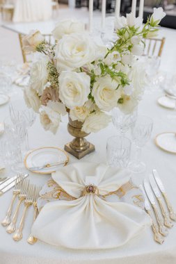 Düğün masasının klasik tabaklar, yaldızlı gümüş takımlar, klasik yüzüklü peçete, kristal ve gül buketli misafir yerinin dikey görüntüsü. Kutlamaların organizasyonu. 