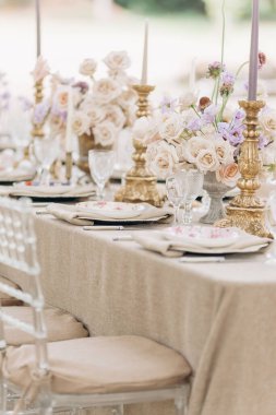 Görkemli ağaçların altındaki eski bir parkta eski şamdanlar, gül buketleri ve kaliteli şamdanlarla bej renkte bir düğün masasının muhteşem klasik dekoruyla yakın plan çekimi.. 