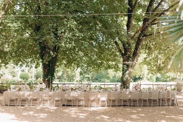 Long wedding table in natural colors with bouquets of flowers in old garden under tall trees. Hotel restaurant business. Wedding floristry design. Organization of festive events. Rest and pleasure.