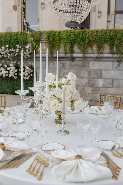 Wedding inpalace style.White wedding table with luxurious setting, bouquet of roses in vintage vase, under an crystal chandelier on terrace of old building. Organization registration of weddings.