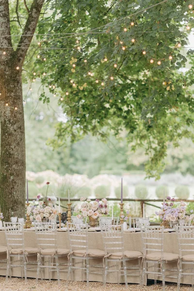 Long wedding table with magnificent luxurious serving with bouquets of roses, vintage candlesticks in old park under majestic trees. Organization and registration of weddings. Special events.