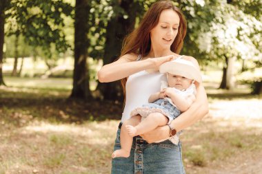 Bekar anne. Kahverengi saçlı, uzun saçlı, yaz kıyafetleri içinde gülümseyen bir kadın. Doğada yaz gününde küçük kızı kollarında. Yeni doğan bakımı. Sağlık sigortası. Çocuk koruması.