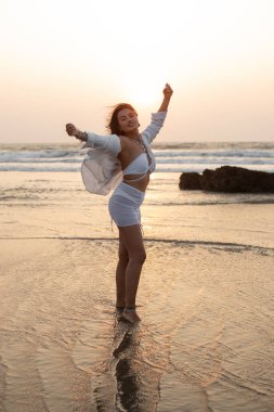 Uzun siyah saçlı, beyaz mayo, gömlek giyen, gün batımında sahile yakın duran, elini kaldıran ve tatilin tadını çıkaran genç Asyalı muhteşem kadının yan görüntüsü. Güzellik, seyahat, okyanus.