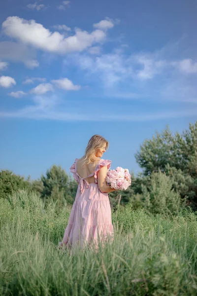 Summer mood. Unrecognizable woman in open summer sundress stands in nature with luxurious bouquet of peonies with her back to camera. Femininity and beauty. Natural cosmetics, skin care. Vertical.