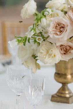 Antika bronz bardakta ve beyaz masa örtüsünün üzerinde duran kristal bardaklarda duran lüks güllerden oluşan bir buket yakın plan. Düğün için. Festival etkinliklerinin dekorasyonu. Beyaz tonlarda düğün.