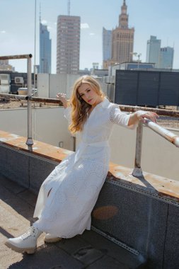 Uzun dalgalı saçları olan çekici sarışın yaz günü binanın çatısında şehrin arka planına karşı poz veriyor. Güzel kadın tasvirleri. Yaşam tarzı. Turizm ve seyahat.