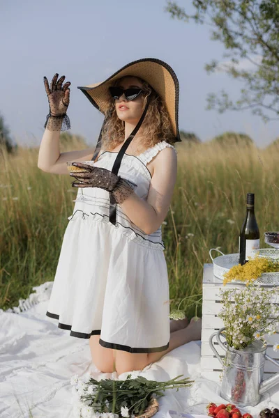 Stock image Picnic in nature on summer day. Charming blonde in white summer dress,elegant hat is drinking champagne, kneeling on white bedspread with fruits, flowers. Romantic mood. Freedom and relaxation. 