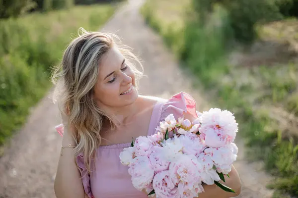 Beautiful portrait of happy woman in nature with luxurious bouquet of peonies on summer day against background of country road. Femininity and beauty. Positive emotions. Romantic mood.