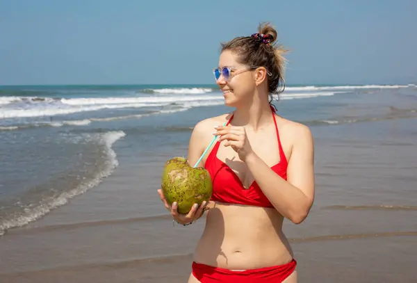 Portrait of young wonderful woman with dark hair wearing red swimsuit, sunglasses standing in water near beach at sunset, holding coconut with straw, enjoying sunshine. Beauty, travelling, ocean.