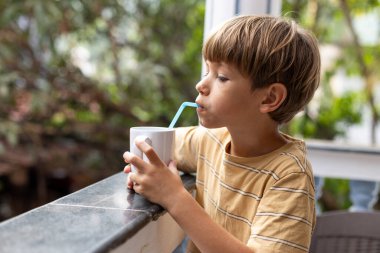 6-7 yaşındaki çocuk, yaz günü terasta oturarak meyve suyu, içecek ve süt içmeyi seviyor. Mutlu bir çocukluk. Yaz tatili. Çocukların sağlığının korunması. 