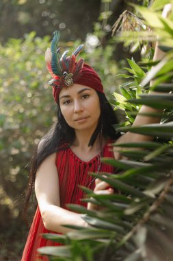 Egzotik kıyafetli güzel kahverengi gözlü esmerin dikey portresi, yaz sabahı tüylü türban, parlak tropikal yeşillikler arasında. Doğayla bütünlük. Esrarengiz. Barış gevşemesi.