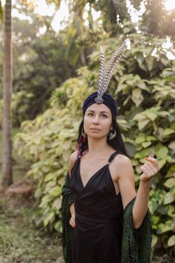 Sarık desenli, siyah elbiseli, doğada duran tüylerle süslenmiş güzel, genç esmer bir kızın portresi. Ruhanilik, ruhani uygulamalar. Dünyayla uyum içinde, kendini ara. 