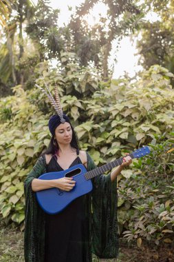 Ruhsal gelişim. Sarıklı, uzun saçlı, tüylerle süslenmiş esmer kadın yaz günü elinde mavi gitarla ormanın arka planında duruyor. Harmony, kendini ara.. 