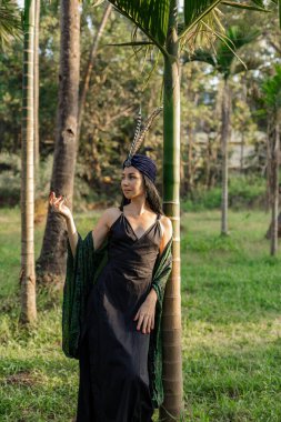 Ağaçların gövdeleri arasında, yaz günü tüylerle süslenmiş türbanlı esmer esmerin tam boy portresi. Ruhani uygulamalar. Harmony, kendini arama. Doğayla bütünlük. Özerklik. 