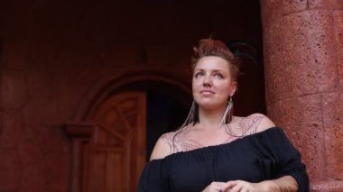 Vücudunda dövmeleri olan çekici bir kadın, orjinal tüy süslemeleri gülümsüyor ve antik binadaki hayattan zevk alıyor. Turizm ve seyahat. Kendine güvenen kadınlar. Mutluluk ve sükunet. 4k görüntü