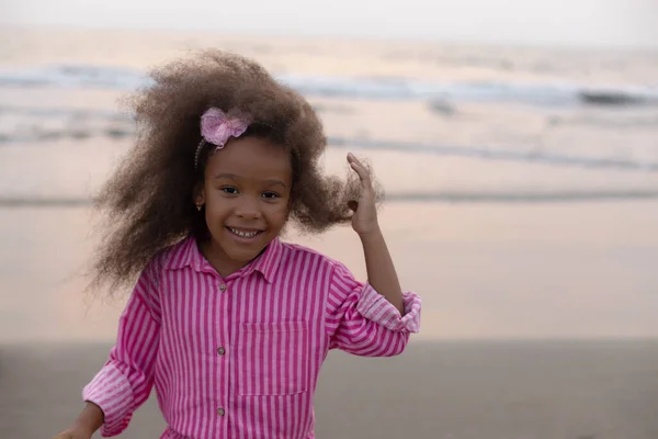 Portrait of charming happy African-American girl in pink shirt on sandy beach, against background of sea, ocean. Happy childhood. Positive childrens emotions. Summer holidays. Recreation travel.