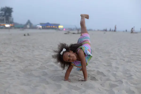 6-7 years old African-American girl does gymnastic somersault on seashore. Girl is gymnast. Child plays sports on beach. Harmonious development of children. Childrens holidays. Happy childhood.