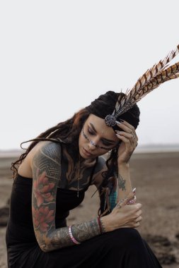 Kafasında tüyleri ve dövmeleri olan bir kadın sahilde oturuyor. Yüksek kalite