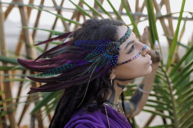 Tüylü saç bandı olan bir kadın palmiye ağacının önünde duruyor, kirpikleri mavi gökyüzüne çarpıyor.