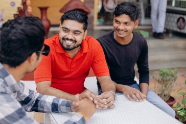 Üç arkadaş açık bir kafede sohbet ediyor, güneşli gökyüzünün altında birbirleriyle eğleniyorlar.