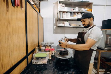 Modern bir kafe mutfağında, çeşitli malzemelerle çevrili bir barista moda bir ortamda kahve pişirirken görülüyor.