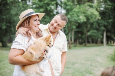 Bir adam ve bir kadın ağaçlar ve bitkilerle çevrili bir parkta küçük bir köpeği mutlu bir şekilde tutuyorlar. Gülümsüyorlar ve doğada boş vakitlerin tadını çıkarıyorlar.