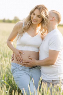 Bir adam, bitkiler ve doğa ile çevrili çimenli bir arazide hamile kadınların karnını öperken gülümsüyor. Bu jest, içinde büyüyen hayata duyduğu sevgi ve mutluluğu gösteriyor.