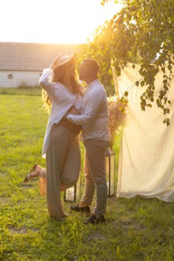 Bir çift, açık mavi bir gökyüzünün altında, uzun otlar ve ağaçlarla çevrili bir çayırda kucaklaşıyor. Kadın dalgalı bir elbise giyiyor ve ikisi de mutlu görünüyor.