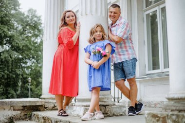 Mutlu bir aile, boş zamanlarında binanın merdivenlerinde fotoğraf çektirmek için gülümsüyor. Bacakları çapraz ve belleri rahat, arka planda elektrikli mavi bir ağaç var.