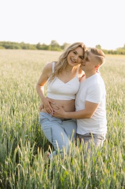 Bebek bekleyen bir çift buğday tarlasında oturuyorlar, otların ve çayırların doğal manzarasıyla çevrili. Gökyüzü açık. Huzurlu ortamın tadını çıkarırken onları mutlu ediyor.