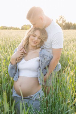 Mutlu bir adam çimenli bir arazide, uzun otlarla çevrili hamile bir kadını kucaklıyor ve huzurlu doğal manzaranın tadını çıkarıyor.