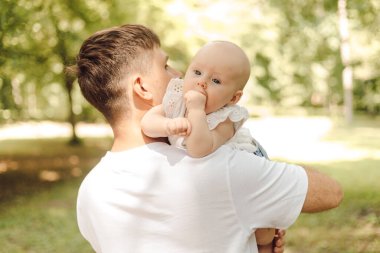 Bir adam yeşil çimenler ve bitkilerle çevrili parkta otururken mutlu bir şekilde bir bebeği kollarında tutuyor. Doğada birlikte vakit geçirmekten hoşlanırlar.