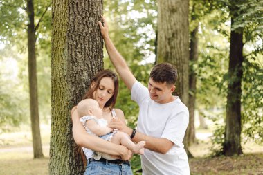 Bir çift, ağaçlar ve çimlerle çevrili bir bebeği kucağında tutarak parkta boş vakitlerin tadını çıkarıyor. Doğal ortamda mutlu ve memnun görünüyorlar.