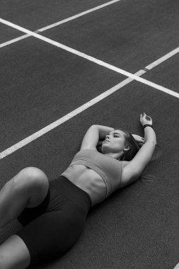 Tenis kortunun zemininde eli ve bacağı uzatılmış, dizleri ve dirseği yere değmiş bir kadın yatıyor. Spor kıyafetleri giyiyor.
