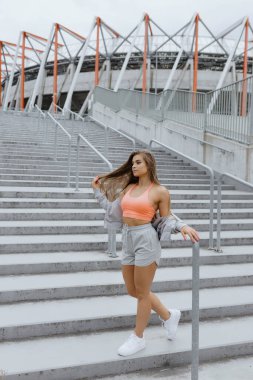 Güneş gözlüklü bir kadın stadyumun önündeki merdivenlerde duruyor. Eğlenceli bir etkinliğe hazır, belini ve kalçasını göstererek gezintiye çıkıyor.