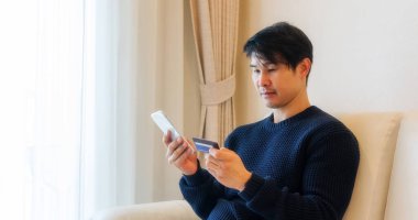 Bir odada akıllı telefon ve kredi kartı kullanan Asyalı bir adam..