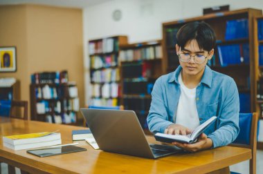 Erkek üniversite öğrencileri dizüstü bilgisayarlar üzerinde çalışıyor ve çalışmak için kitaplar arıyor, raporlar hazırlıyor, üniversite odasında yararlı bilgiler buluyor. Okuma, öğrenme ve zeka kavramı.