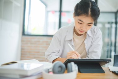 Genç Asyalı üniversite öğrencisi rapor hazırlıyor, kafede tablet çiziyor. İnternette iletişim ve araştırma.