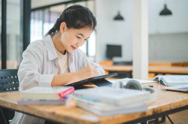 Genç Asyalı üniversite öğrencisi rapor hazırlıyor, kafede tablet çiziyor. İnternette iletişim ve araştırma.