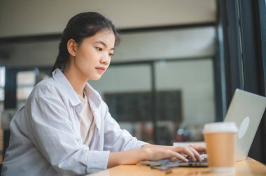 Genç Asyalı üniversite öğrencisi kafede dizüstü bilgisayarda çalışıyor ve öğreniyor. İnternette bilgi aranıyor.