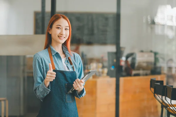 Smiling Asian coffee shop owner standing in front of cashier counter with tablet ready to take menu orders.welcome customers who come in to place orders in the cafe.