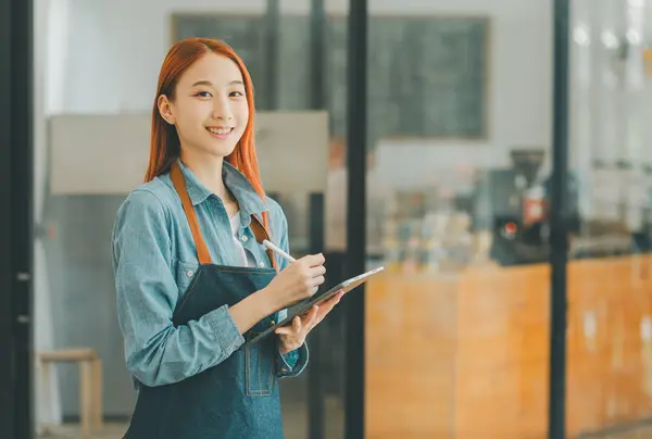 Smiling Asian coffee shop owner standing in front of cashier counter with tablet ready to take menu orders.welcome customers who come in to place orders in the cafe.