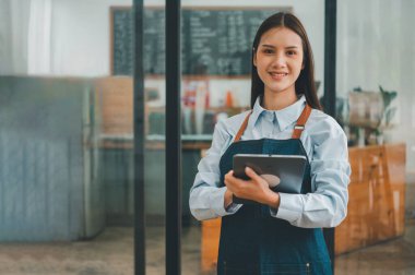 Gülümseyen Asyalı kahve dükkanı sahibi elinde tabletle kasiyerin önünde menü siparişlerini almaya hazır bekliyor. Kafeye sipariş vermek için gelen müşterileri hoş karşılar..