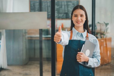 Gülümseyen Asyalı kahve dükkanı sahibi elinde tabletle kasiyerin önünde menü siparişlerini almaya hazır bekliyor. Kafeye sipariş vermek için gelen müşterileri hoş karşılar..