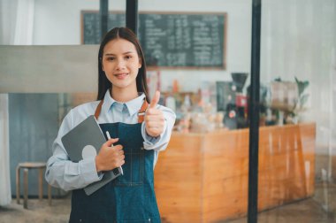 Gülümseyen Asyalı kahve dükkanı sahibi elinde tabletle kasiyerin önünde menü siparişlerini almaya hazır bekliyor. Kafeye sipariş vermek için gelen müşterileri hoş karşılar..