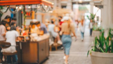 Kalabalık bir açık hava pazarının görüntüsü bulanık. Etrafta yürüyen ve masalarda oturan insanlar var. Atmosfer canlı ve hareketli.