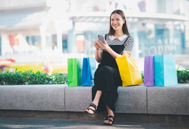Elinde cep telefonu ve önünde birkaç alışveriş poşeti olan bir kadın bankta oturuyor.