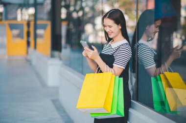 Bir kadın elinde cep telefonu ve iki alışveriş çantasıyla dışarıda bekliyor. Gülümsüyor ve alışveriş deneyiminin tadını çıkarıyor.