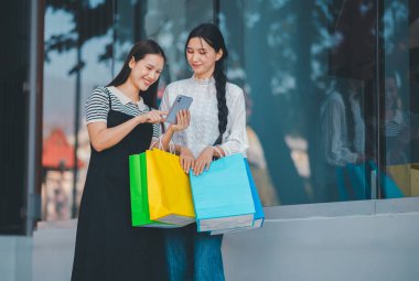 İki kadın bir mağazanın önünde cep telefonlarına bakıyor. Alışveriş çantalarını tutuyorlar ve telefonlarından bir şeyi kontrol ediyor gibi görünüyorlar.